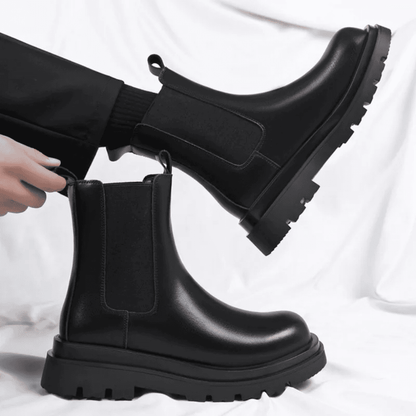 FORCIANO SENTINEL black elevator Chelsea boots shown as a pair, with one worn and one held, highlighting the elastic side panels and premium leather uppers.